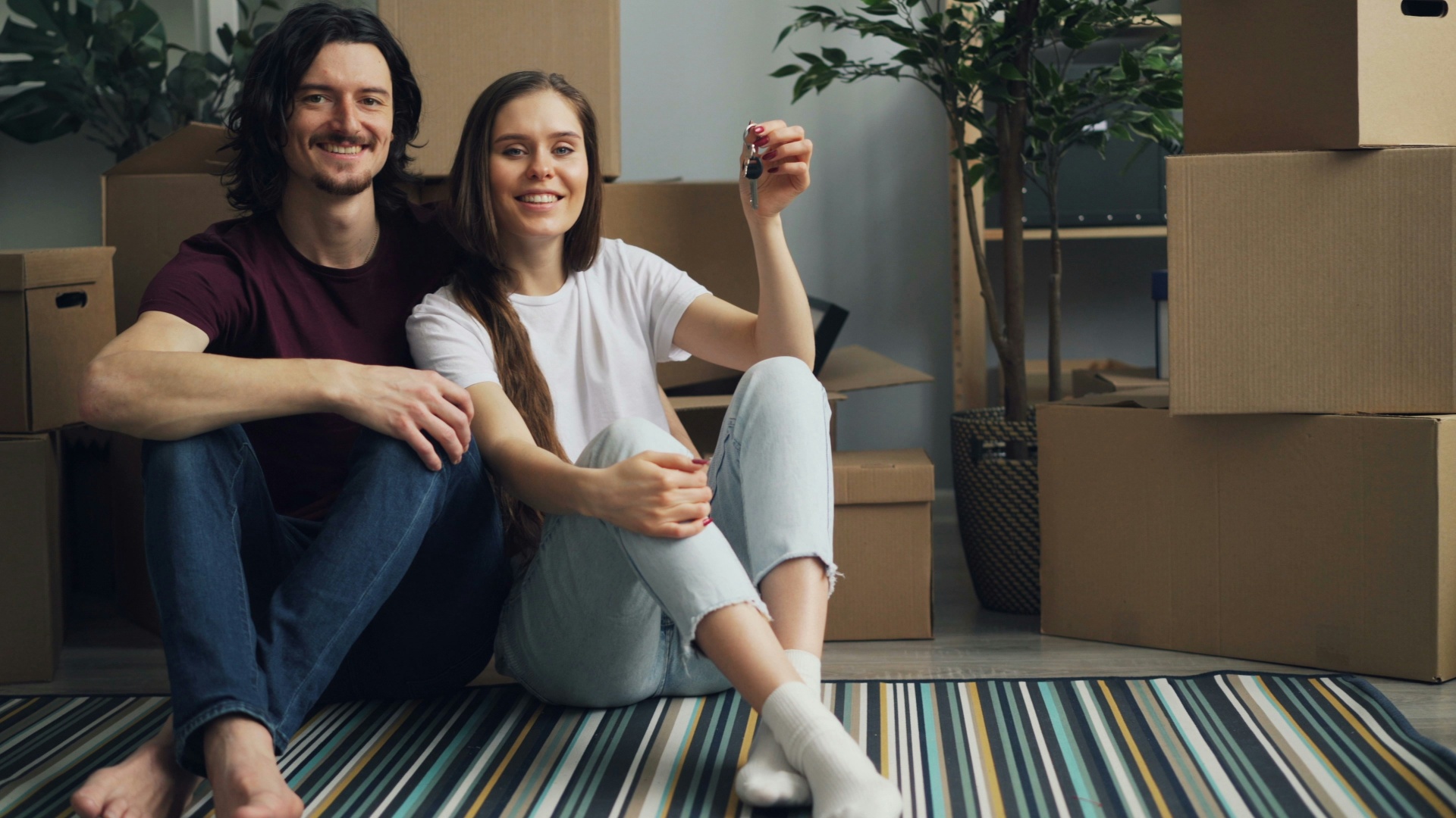 Pareja sonriente con llaves de su nueva vivienda rodeada de cajas de mudanza